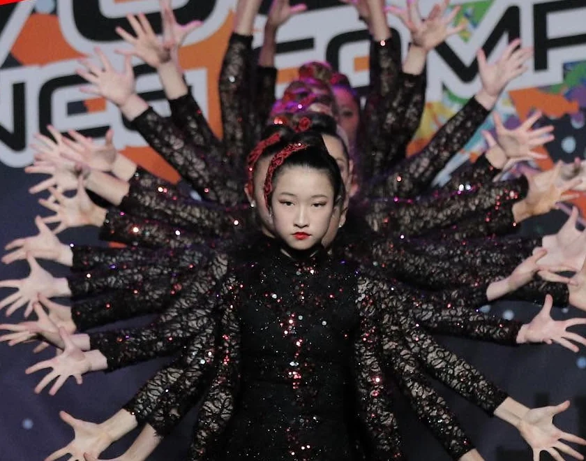 JMDS troupe in black sequins lined up behind one dancer so their arms fan out like a burst